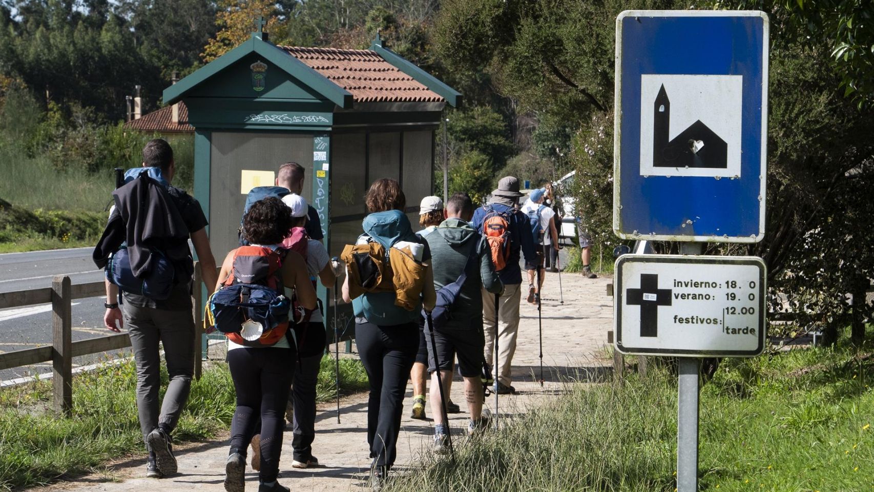 Un grupo de peregrinos pasa frente a un cartel que indica el horario de misas mientras realizan el Camino de Santiago.